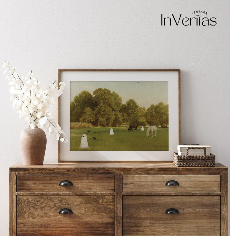 May include: A framed print featuring a pastoral scene with white ghost figures in a green field with horses and trees. The artwork is displayed on a wooden dresser with a vase of white flowers.