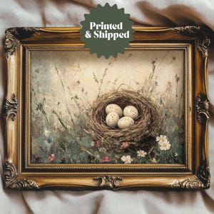 May include: A framed painting of a bird's nest with four white speckled eggs inside. The nest is made of twigs and is surrounded by green leaves and flowers. The painting is set against a light brown background.