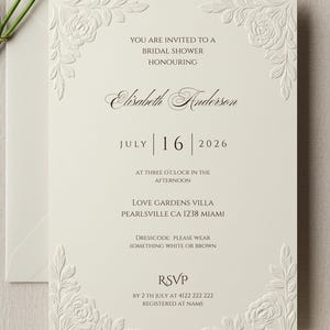 May include: Ivory bridal shower invitation with embossed floral corner designs and text. The invitation includes the date July 16, 2026, and RSVP information. A white rose and envelope are in the background.