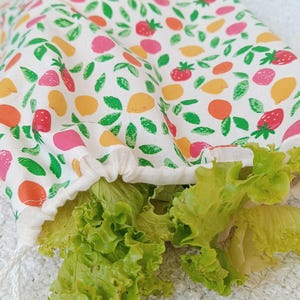 Peut inclure: Un sac à légumes blanc à cordon de serrage rempli de laitue verte. Le sac est orné d'un motif coloré de fruits et de feuilles vertes. Le motif comprend des fraises, des citrons et des oranges.