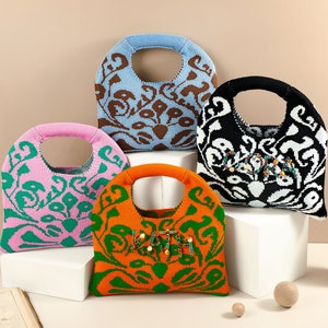 May include: Four crocheted handbags in different colors and patterns. The bags have a rounded shape and a single handle. The bags are displayed on a white surface with a book and wooden balls.