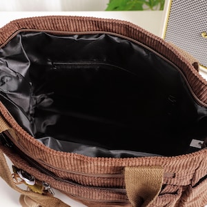 Puede incluir: Vista superior de un bolso tote de pana marr&oacute;n con un interior negro. El bolso tiene un compartimento principal con cremallera y m&uacute;ltiples bolsillos exteriores. Cuenta con asas de lona marr&oacute;n y herrajes plateados.