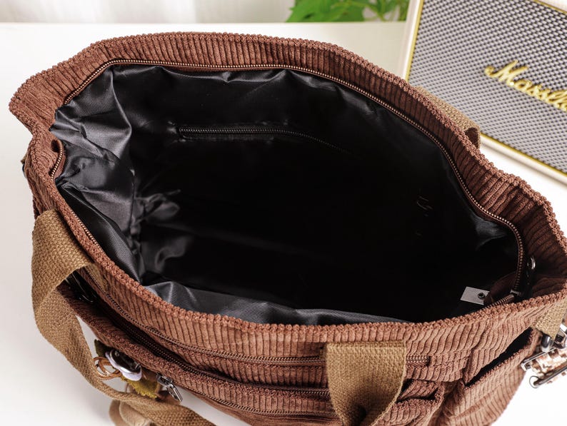 Puede incluir: Vista superior de un bolso tote de pana marr&oacute;n con un interior negro. El bolso tiene un compartimento principal con cremallera y m&uacute;ltiples bolsillos exteriores. Cuenta con asas de lona marr&oacute;n y herrajes plateados.