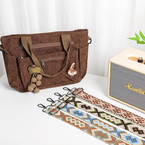 Puede incluir: Un bolso tote de pana marr&oacute;n con m&uacute;ltiples bolsillos y un asa superior. El bolso tiene un amuleto decorativo de uva y p&aacute;jaro. Tres correas para el hombro estampadas con cierres met&aacute;licos se muestran frente al bolso.