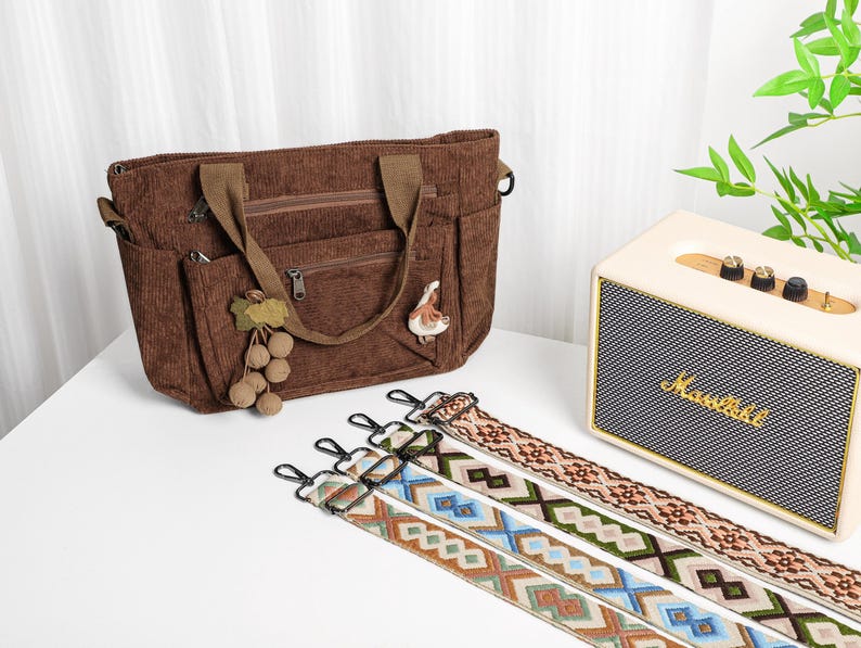 Puede incluir: Un bolso tote de pana marr&oacute;n con m&uacute;ltiples bolsillos y un asa superior. El bolso tiene un amuleto decorativo de uva y p&aacute;jaro. Tres correas para el hombro estampadas con cierres met&aacute;licos se muestran frente al bolso.