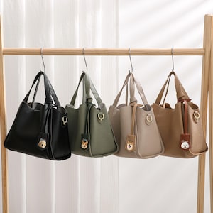 Può includere: Quattro borse in pelle nera, verde oliva, talpa e marrone chiaro sono appese a un supporto in legno. Ogni borsa ha un manico superiore, una tracolla staccabile e un ciondolo decorativo. Le borse sono esposte su uno sfondo bianco.