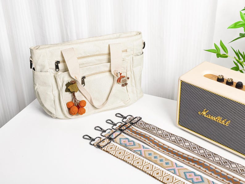 Puede incluir: Bolso tote de pana color crema con m&uacute;ltiples bolsillos y cremalleras, adornado con abalorios decorativos. Incluye varias correas para el hombro estampadas con cierres met&aacute;licos. Un altavoz crema y negro est&aacute; en el fondo.