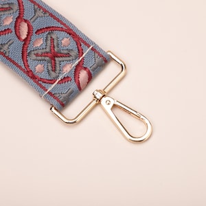 May include: A close-up of a blue and red patterned fabric strap with a gold metal clasp. The strap is attached to a gold metal hook.