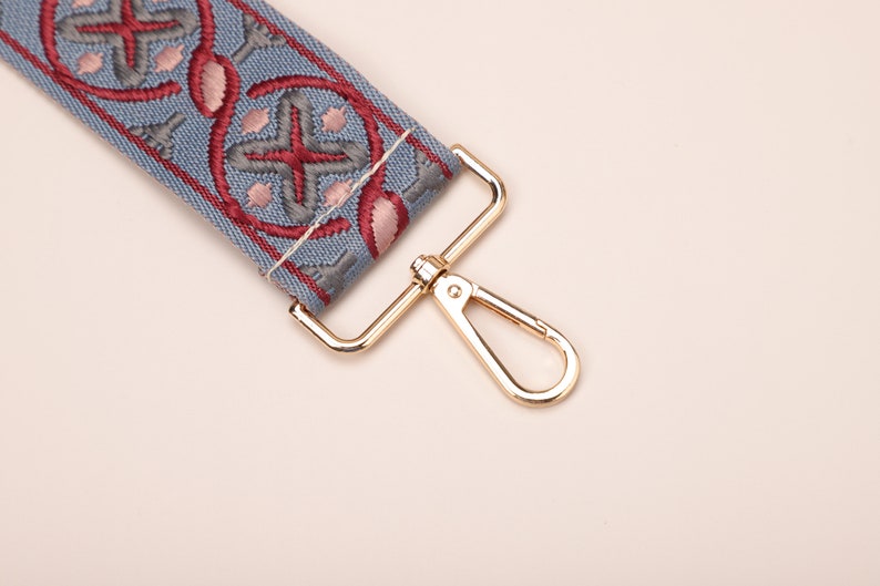 May include: A close-up of a blue and red patterned fabric strap with a gold metal clasp. The strap is attached to a gold metal hook.