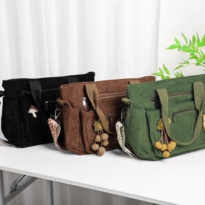 Puede incluir: Tres bolsos tote de pana en negro, marr&oacute;n y verde, cada uno con una correa para el hombro y m&uacute;ltiples bolsillos. La bolsa negra tiene un amuleto de hongo, la bolsa marr&oacute;n tiene una borla y la bolsa verde tiene un parche floral.