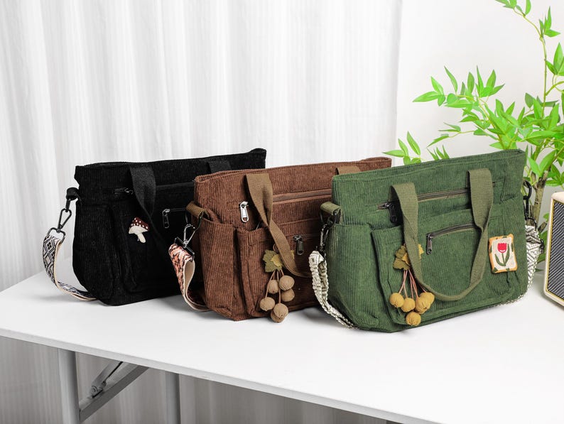 Puede incluir: Tres bolsos tote de pana en negro, marr&oacute;n y verde, cada uno con una correa para el hombro y m&uacute;ltiples bolsillos. La bolsa negra tiene un amuleto de hongo, la bolsa marr&oacute;n tiene una borla y la bolsa verde tiene un parche floral.
