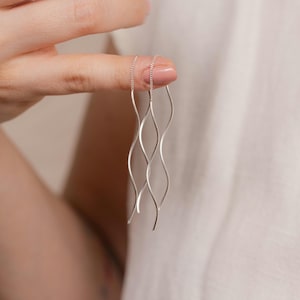 Può includere: Un paio di orecchini pendenti argentati ondulati. Gli orecchini sono tenuti da un dito, mostrando il loro design delicato e fluido. Gli orecchini hanno una catena nella parte superiore e una forma curva e allungata.