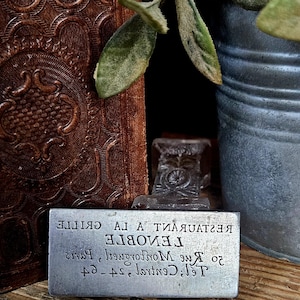 Könnte beinhalten: Ein rechteckiger Metallstempel mit spiegelverkehrt eingraviertem Text, der RESTAURANT A LA GRILLE, LENOBLE, 20 Rue Montorgueil, Paris, Tel: Central 24-64 lautet. Der Stempel hat einen dunklen, verzierten Griff und steht aufrecht für gute Lesbarkeit.