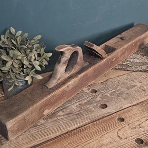 May include: A long, vintage wooden hand plane featuring a dark brown finish, a curved handle, and a metal blade assembly set into the top. The tool has a rectangular body with visible wood grain and signs of age, showcasing a classic carpentry design.