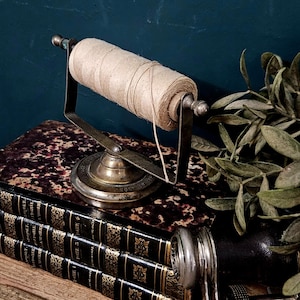 Op de afbeelding: Een vintage-stijl messing touwhouder met een cilindrische spoel van natuurlijk beige touw, vastgehouden door een metalen frame op een ronde voet. De houder staat bovenop een stapel van drie zwart lederen boeken met gouden details.