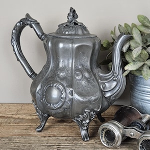 Op de afbeelding: Een metallic grijze tinnen theepot met een sierlijk reliëfpatroon van bloemen, een gebogen handvat, een decoratief deksel met een knop en vier krulpoten. Het lichaam van de theepot toont een centraal rond medaillon en gedetailleerde reliëfs.