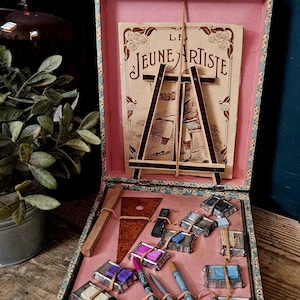 Op de afbeelding: Een vintage kunstbenodigdhedendoos met een bloemmotief aan de buitenkant en een roze binnenkant. Het bevat een kleine houten ezel, een papieren afdruk met het label LE JEUNE ARTISTE, een houten liniaal, een driehoekig hulpmiddel en diverse kleine verfblokjes.