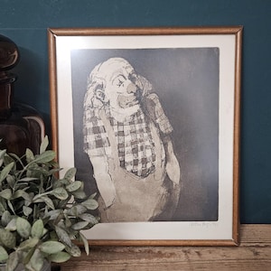 May include: A framed black and white line drawing of a clown wearing a checkered shirt and overalls. The artwork features a signature and the year 1975 in the bottom right corner. The print is encased in a simple, thin, light brown wooden frame.