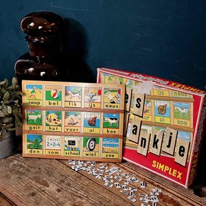 Puede incluir: Un juego educativo vintage Simplex que incluye un tablero de madera con quince tarjetas ilustradas que muestran palabras en neerlandés como jaap, bal, fik, ijs, neus, geit, hut, vuur, ui, koe, den, som, school, wiel y zee, junto a una caja roja y letras sueltas.