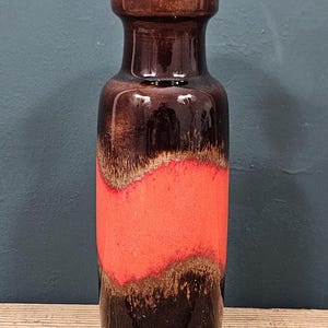 Op de afbeelding: Een hoge keramische vaas met een donkerbruine boven- en onderkant, met een levendige rode band in het midden. De vaas heeft een glanzende afwerking en een uniek retro-ontwerp, geschikt voor interieurdecoratie.