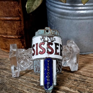 Puede incluir: Una placa de metal vintage para bicicleta con un acabado desgastado y oxidado. La placa muestra el texto VELOS en negro, SISSEN en letras rojas grandes y VILVOORDE verticalmente en un panel de esmalte azul, todo sobre un fondo blanco.