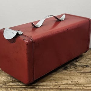 Op de afbeelding: Een rechthoekige opbergdoos van rood metaal met afgeronde hoeken en een zilverkleurig metalen handvat aan de bovenkant. De doos heeft een scharnierend deksel met een zilverkleurige metalen sluiting aan de zijkant en vertoont tekenen van slijtage.