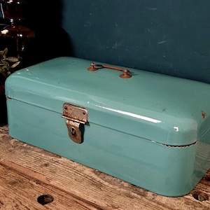 May include: A rectangular, light teal enameled metal bread box with rounded corners. It features a top-mounted metal handle and a front-facing metal latch mechanism, both showing signs of rust and wear on the surface of the container.