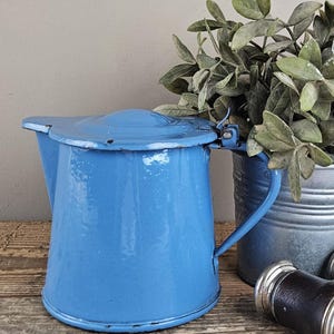 Op de afbeelding: Een felblauwe geëmailleerde metalen kan met een scharnierend deksel en een gebogen handvat. De kan heeft een taps toelopend cilindrisch lichaam met een brede basis en een kleine schenktuit. Het oppervlak vertoont lichte gebruikssporen.