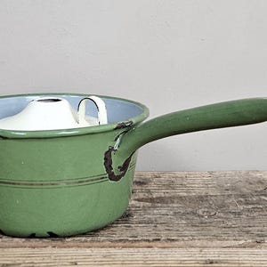Op de afbeelding: Een groene geëmailleerde metalen steelpan met een lange steel en een schenktuit. De binnenkant is lichtblauw en de buitenkant heeft donkerbruine, versleten randen. In de pan ligt een wit deksel met een klein handvat en een opening.