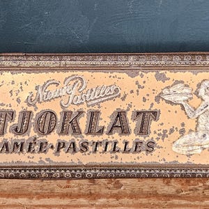 Op de afbeelding: Een rechthoekig vintage metalen blik met een verweerde goud- en bruinkleurige afwerking. Het deksel bevat de tekst "Nieuwe Pastilles", "TJOKLAT" en "CAMEE PASTILLES" naast een afbeelding van een knielende figuur die een schaal vasthoudt.