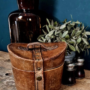 Può includere: Una scatola portaoggetti in pelle marrone in stile vintage con coperchio curvo, dotata di una cinghia verticale centrale fissata con borchie in metallo color ottone e una maniglia abbinata. La pelle ha una finitura usurata e strutturata.