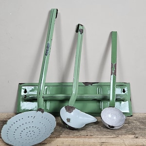 Op de afbeelding: Een set van drie lichtgroene geëmailleerde keukengerei met lange stelen en een bijpassend rechthoekig wandrek. De set bevat een grote schuimspaan, een middelgrote soeplepel en een kleine soeplepel, alle met zichtbare gebruikssporen.