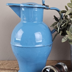 Op de afbeelding: Een lichtblauwe emaille kan met een rond lichaam, een uitlopende voet, een gebogen handvat en een scharnierend deksel. Het metalen oppervlak vertoont gebruikssporen met kleine beschadigingen en donkere vlekken langs de randen.