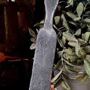 May include: A hand-forged iron spatula with a long, flat, rectangular blade and a tapered handle featuring a circular hole at the top. The metal surface has a dark, textured, matte finish consistent with traditional blacksmithing techniques.