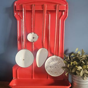 Pode incluir: Um suporte de parede para utensílios de cozinha em metal esmaltado vermelho brilhante, com uma bandeja de gotejamento inferior e uma barra horizontal. Quatro utensílios de cozinha brancos esmaltados com cabos longos estão pendurados, incluindo uma concha e uma escumadeira.