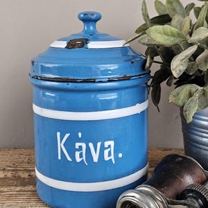 Pode incluir: Um recipiente cilíndrico de esmalte azul com tampa, apresentando duas faixas brancas horizontais e a palavra Kava. impressa em letras brancas na frente. O recipiente mostra sinais de desgaste com manchas de ferrugem castanho-escuras.
