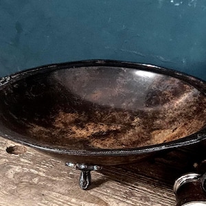 Könnte beinhalten: Eine ovale Schale aus dunklem Metall mit einer verwitterten, fleckigen braunen und schwarzen Patina. Die Schale verfügt über zwei verzierte, strukturierte Griffe an den gegenüberliegenden Seiten und steht auf drei kleinen, dekorativen Füßen.