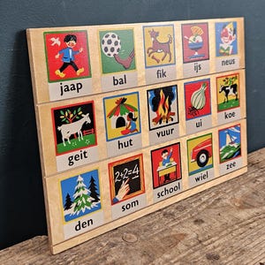 Op de afbeelding: Houten educatief bord met kleurrijke illustraties en Nederlandse woorden. Elke vierkant toont een afbeelding en een woord, waaronder "jaap", "bal", "fik", "ijs", "neus", "geit", "hut", "vuur", "ui", "koe", "som", "school", "wiel", "zee" en "den".