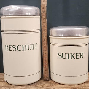 Puede incluir: Dos recipientes cilíndricos de color crema con tapas plateadas y texto en verde. El recipiente más alto tiene la etiqueta BESCHUIT y el más bajo SUIKER. Una regla plegable de madera colocada entre ellos muestra una altura de 23 cm.