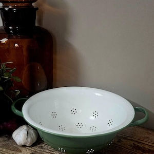 May include: A round, two-handled colander with a sage green exterior and a smooth white interior. The bowl features a series of small, evenly spaced drainage holes arranged in clusters across the base and sides of the basin.