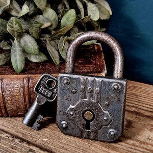 Puede incluir: Un candado de metal gris oscuro desgastado con su llave a juego. El candado presenta una cerradura central, remaches decorativos y la palabra "SECURITY" grabada en el frente. La llave tiene el número "122" marcado en su cabeza rectangular.