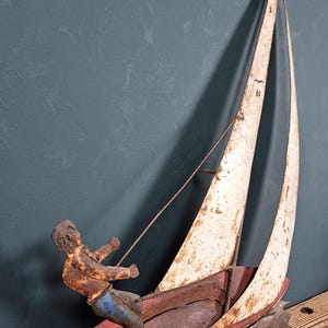 Puede incluir: Una escultura decorativa de velero de metal con una vela blanca desgastada con el número 60, un casco rojo y una figura con camisa marrón y pantalones azules. La base es una pieza de metal texturizada, oscura y dentada con el número 79.