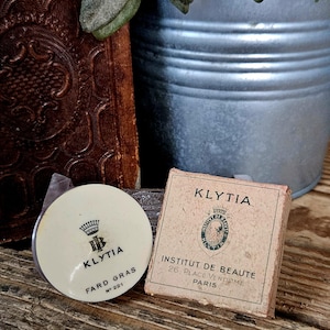 Op de afbeelding: Een ronde Klytia Fard Gras No 221 crèmehouder met een kroonlogo en een vierkante Klytia Institut de Beauté doos met een wapenschild en het adres 26 Place Vendome Paris gedrukt in zwarte inkt op een lichtbeige papieren ondergrond.