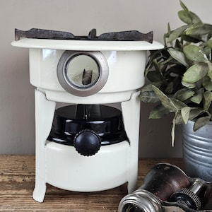 May include: A vintage-style cream-coloured portable stove with a black fuel tank and a central control knob. The unit features a circular glass viewing window on the front, a dark metal burner grate on top, and a sturdy white metal frame base.