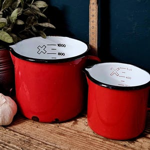 Op de afbeelding: Twee rode emaille maatbekers met een witte binnenkant en zwarte randen. De grotere beker heeft zwarte maatstreepjes tot 1000 ml, terwijl de kleinere beker markeringen tot 500 ml toont. Beide zijn voorzien van een handvat en een schenktuit.