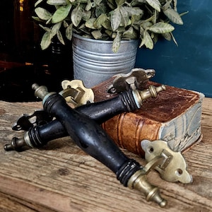May include: Two drawer pulls featuring long, cylindrical black wooden handles with brass-toned metal ends and mounting plates. The handles have a smooth, tapered design with ribbed detailing near the ends, connected to ornate brass-colored hardware.