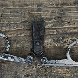 May include: A pair of vintage folding scissors made of dark, weathered metal. The tool features two circular finger loops connected to a central pivot point with short, pointed blades. The metal surface shows signs of oxidation and wear.