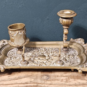 Peut inclure: Un ensemble de bureau en laiton antique avec un plateau décoratif, un porte-stylo et un bougeoir. Le plateau présente un motif floral orné. L'ensemble est en laiton et a un aspect vintage.