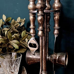 Op de afbeelding: Een zilverkleurige metalen barset op een ronde standaard met een ringhandvat aan de bovenkant. De set bevat een flesopener en een jigger, met versierde, getextureerde handvatten met een verweerde afwerking, staand op een brede, geribbelde voet.