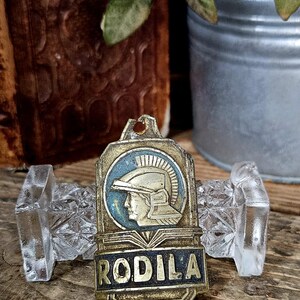 Pode incluir: Uma placa metálica retangular de cor latão com um emblema circular de um perfil de uma figura com capacete num campo azul. Abaixo do emblema, a palavra RODILA está gravada em letras escuras e em negrito. A placa tem orifícios de montagem.